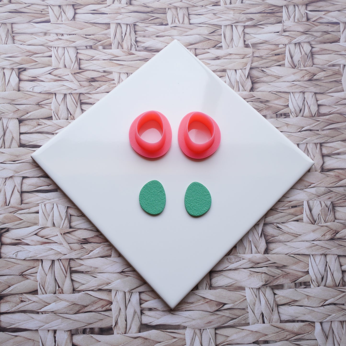 There is a white ceramic tile sitting on top of a straw background. Pictured on this ceramic tile is a Sabi drop-shaped polymer clay pink cutter alongside sage green polymer clay that has been cut out using that cutter.