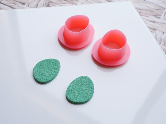There is a white ceramic tile sitting on top of a straw background. Pictured on this ceramic tile is a Sabi drop-shaped polymer clay pink cutter alongside sage green polymer clay that has been cut out using that cutter.