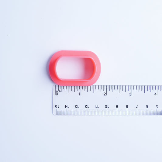 Dog tag-shaped polymer clay cutter 3D printed in pink PLA filament next to clear acrylic ruler to give shoppers reference of where the size options are referencing on the cutter.