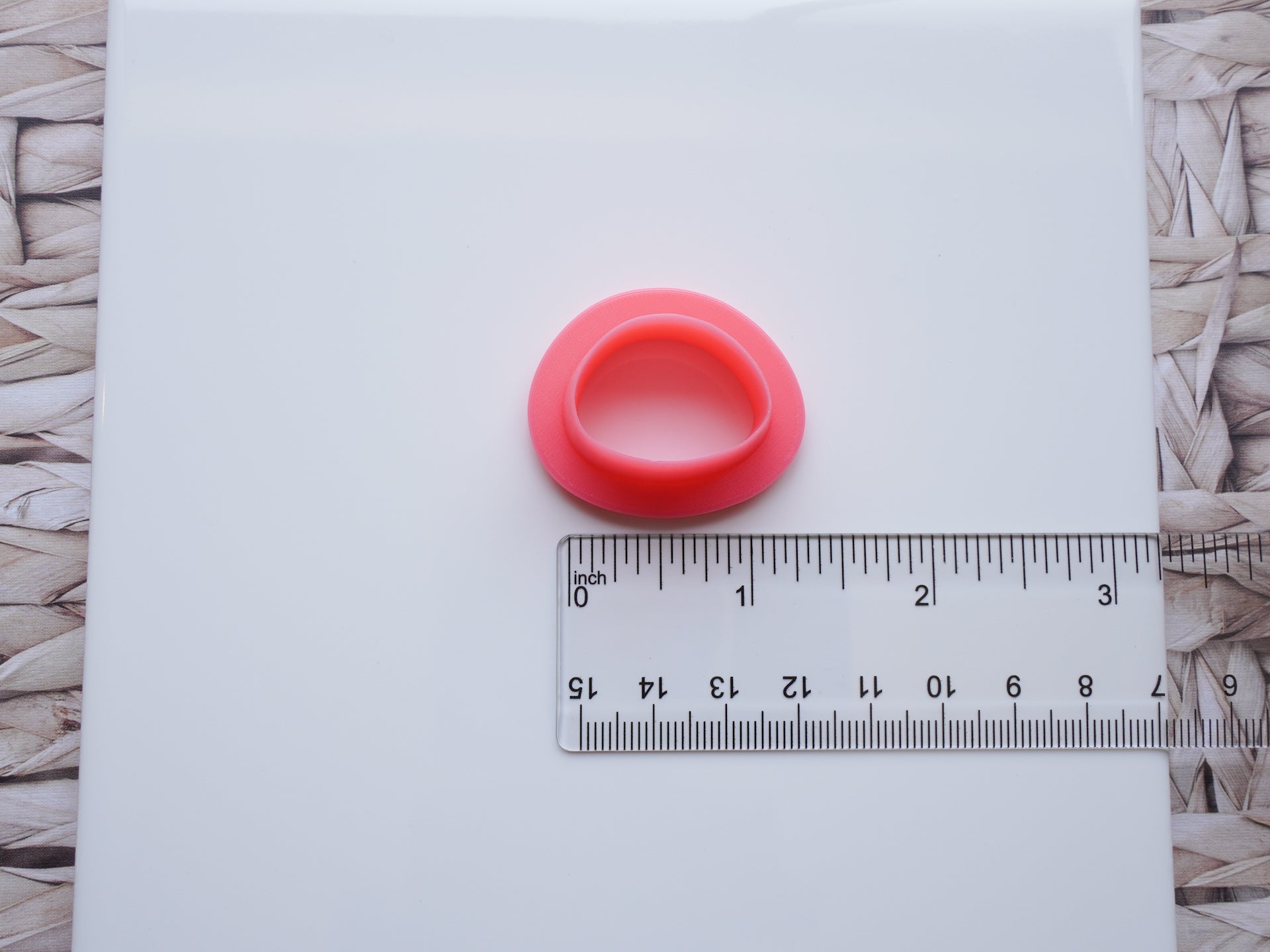 There is a white ceramic tile sitting on top of a straw background. A Sabi drop-shaped polymer clay pink cutter is pictured on this ceramic tile with a clear acrylic ruler.