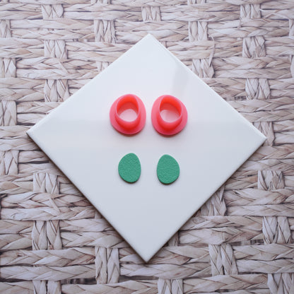 There is a white ceramic tile sitting on top of a straw background. Pictured on this ceramic tile is a Sabi drop-shaped polymer clay pink cutter alongside sage green polymer clay that has been cut out using that cutter.