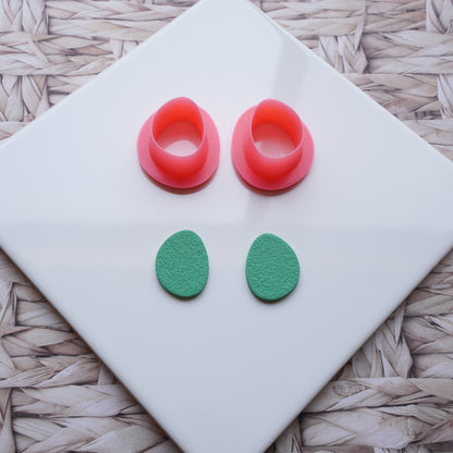 There is a white ceramic tile sitting on top of a straw background. Pictured on this ceramic tile is a Sabi drop-shaped polymer clay pink cutter alongside sage green polymer clay that has been cut out using that cutter.