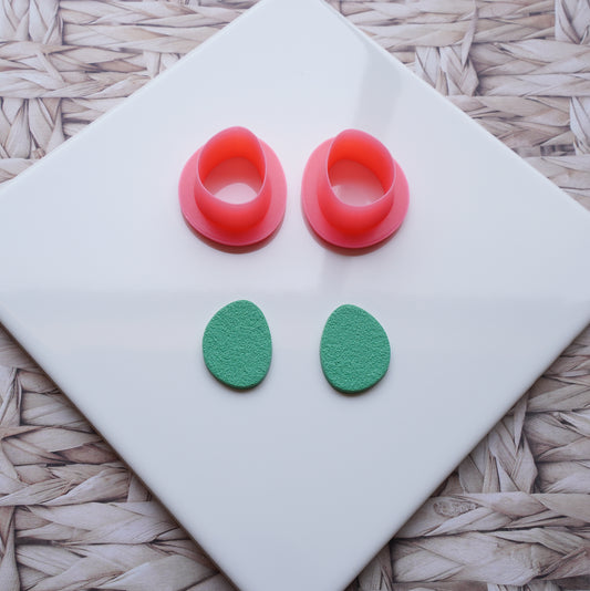 There is a white ceramic tile sitting on top of a straw background. Pictured on this ceramic tile is a Sabi drop-shaped polymer clay pink cutter alongside sage green polymer clay that has been cut out using that cutter.