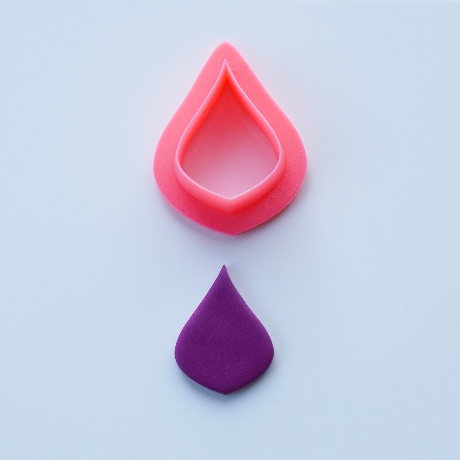 Pink Pear shaped clay cutter is shown above a purple cutout of polymer clay on a white background.